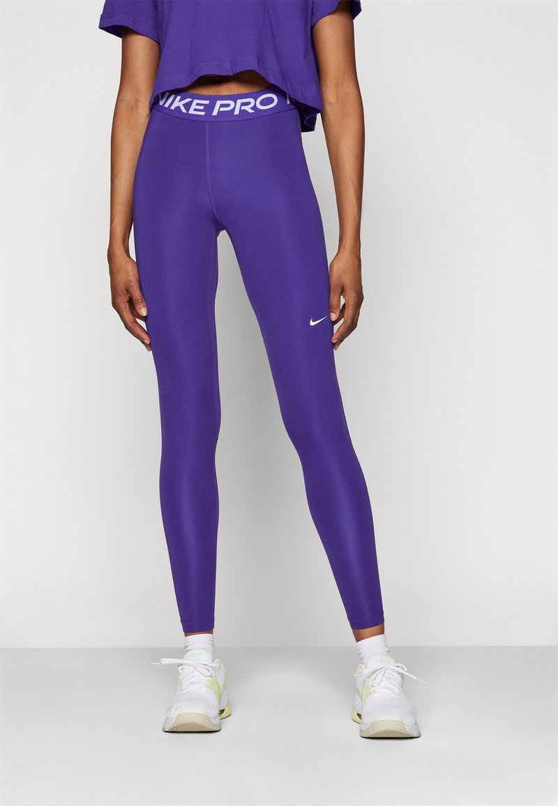 Fioletowe legginsy Nike Pro z szerokim, markowym pasem w talii i małym logo na udzie, w połączeniu z białymi butami sportowymi.