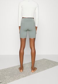 Šedé sportovní šortky s vysokým pasem, zkombinované s bílým dlouhým rukávem, které nosí osoba stojící bosky na texturované podložce.