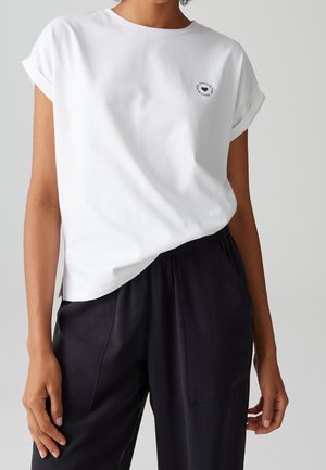 Personne portant un T-shirt blanc à manches courtes avec un petit cœur noir et un texte sur la poitrine, associé à un pantalon noir ample.