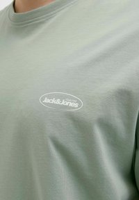 Világoszöld pamut póló, kerek nyakkivágással, rövid ujjakkal, és egy fehér ovális logóval, amely a mellen a "Jack & Jones Club 1990" feliratot viseli.