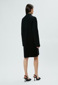 Cappotto nero realizzato in tessuto strutturato, con una vestibilità lunga e ampia, maniche larghe e colletto alto, abbinato a scarpe nere con tacco.