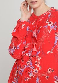 Blouse florale rouge avec des détails froncés. Présente des motifs de fleurs violettes et blanches, des boutons sur le devant et des manches longues. Tissu doux.