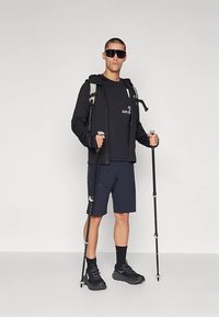 Hombre vestido con ropa de senderismo negra, gafas de sol y mochila, sosteniendo bastones de trekking, de pie frente a un fondo blanco liso.
