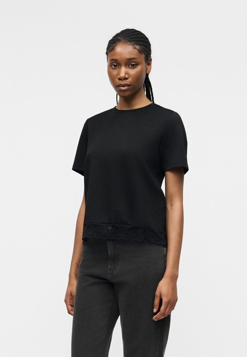 Γυναίκα με πλεγμένα μαλλιά που φοράει μαύρο τοπ με κοντά μανίκια και δαντέλα και σκούρο γκρι τζιν, που στέκεται μπροστά σε ένα απλό φωτεινό φόντο.