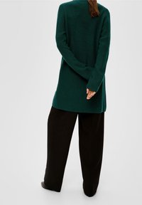 Cardigan lungo di un verde scuro, con una texture a coste, caratterizzato da maniche lunghe, un'apertura frontale e un orlo posteriore leggermente più lungo. Abbinato a pantaloni neri.