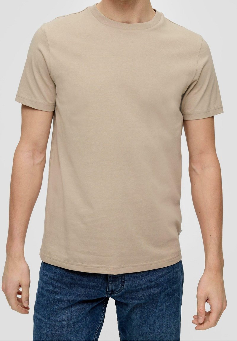 Homme portant un T-shirt beige uni à manches courtes associé à un jean bleu, montré du cou aux hanches sur un fond uni.