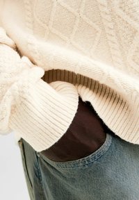 Cremfarbener Zopfmuster-Pullover mit gerippten Bündchen und strukturiertem Muster, der teilweise dunkle Hosen verdeckt. Weicher Stoff mit definierten Nähdetails.