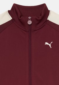Burgundowa kurtka sportowa z prążkowanym kołnierzem, pełnym zapięciem na zamek i białymi akcentami po bokach. Haftowane logo na klatce piersiowej. Miękka tekstura materiału.