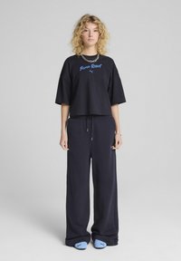 Sort cropped t-shirt med blåt "Puma Resort" tekst, parret med løse navy sweatpants og lyseblå slippers. Enkel, afslappet design.