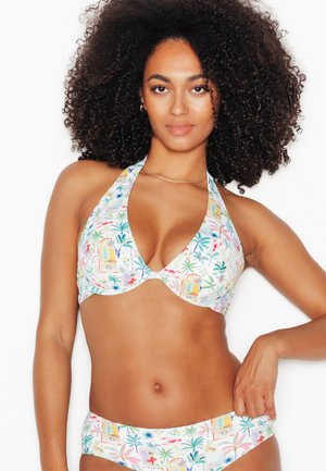Donna con capelli ricci che indossa un bikini halter bianco con stampa tropicale, che posa con una mano sul fianco su uno sfondo semplice.