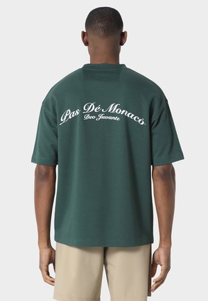 Mann iført mørkegrønn kortermet skjorte med hvit tekst "Pas Dé Monaco Deo Juvante" på ryggen, kombinert med beige shorts.