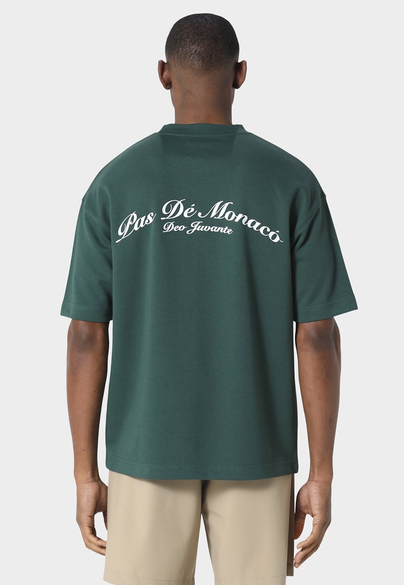 Hombre con camisa de manga corta de color verde oscuro con texto blanco "Pas Dé Monaco Deo Juvante" en la espalda, combinado con pantalones cortos beige.