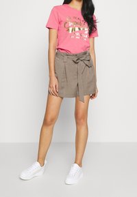 Roze T-shirt met korte mouwen en gouden folie-tekst, gecombineerd met grijze shorts met ceintuur. Witte sneakers maken de outfit compleet en tonen een casual stijl.