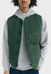 Grüne gesteppte Weste mit einem Knopfverschluss, ausgestattet mit großen Taschen und einer glatten Textur, die über einem grauen Kapuzenshirt getragen wird.
