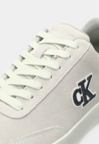 Αθλητικό παπούτσι από γκρι σουέντ με υφή, διαθέτει επίπεδα λευκά κορδόνια και σκούρο μπλε λογότυπο «cK» στο πλάι. Στρογγυλερό σχέδιο μύτης.