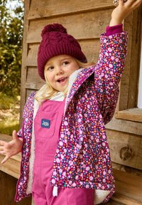 Bunte Jacke mit Blumenmuster und einem mit Fell gefütterten Kapuze, kombiniert mit einem pinken Overall. Strickmütze in Bordeaux mit einem Bommel oben.