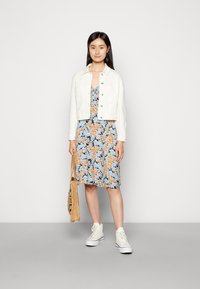 Casaco de ganga branco sobre um vestido floral com padrões azuis, laranjas e brancos. Ténis brancos e uma bolsa de palha completam o visual.