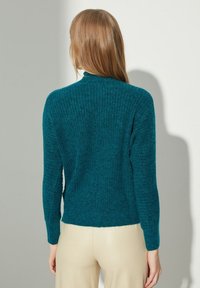 Tyrkysový pletený svetr s ribbovanou texturou a dlouhými rukávy. Má vysoký límec a mírně volný střih. Minimalistický design.