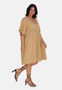 Robe en coton beige texturé avec un décolleté en V et des manches courtes, présentant une coupe ample qui arrive au milieu de la cuisse. Associée à des talons noirs à lanières.