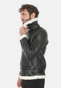 Giacca di pelle nera con colletto alto e rifiniture in shearling bianco. Presenta cerniere laterali, una vestibilità slim e un materiale liscio e testurizzato.