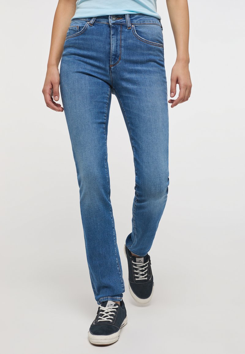 Kék farmernadrág, slim fit fazonnal, diszkrét kopással, öt zsebbel és kontrasztos varrással, fekete sportcipőkkel párosítva.