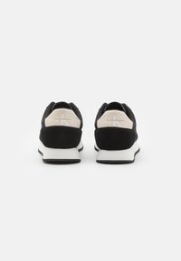 Zapatillas deportivas negras con parte superior de gamuza y malla, suela blanca y detalle de marca en el talón en color crema claro. Textura suave, forma redondeada.