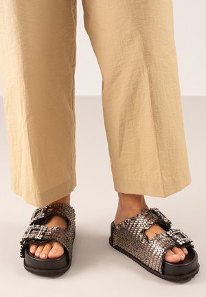 Sandalias metálicas trenzadas con una suela negra gruesa, que presentan hebillas decorativas y un diseño de punta abierta. Superficie texturizada con detalles trenzados.