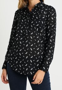 Chemise noire à manches longues avec col, présentant un motif de plumes blanches, une poche poitrine unique et une coupe décontractée. Tissu en coton, légèrement texturé.