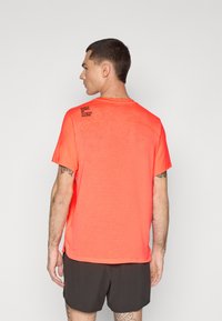 Lys orange atletisk t-shirt med korte ærmer, mesh-struktur og trykt sort tekst på venstre skulder. Båret med mørke shorts.