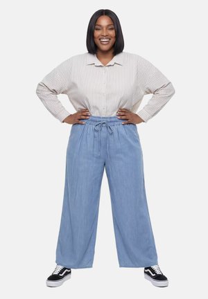 Uśmiechnięta kobieta o włosach do ramion stoi z rękami na biodrach, ubrana w paskowaną koszulę zapinaną na guziki, jasnoniebieskie szerokie spodnie i czarne trampki.