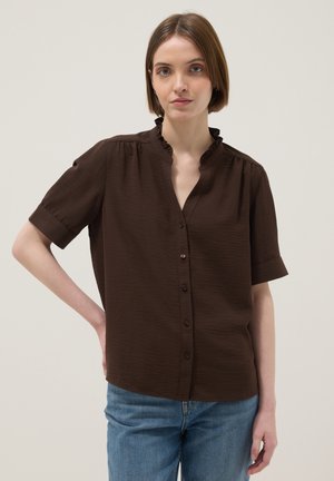 Vrouw met kort bruin haar die een donkergestreept blouse met knopen en een gerimpelde kraag draagt, gecombineerd met een blauwe spijkerbroek, staand tegen een effen achtergrond.