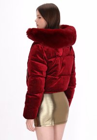 Chaqueta puffer recortada de pana roja con capucha de piel sintética, que presenta una textura acolchada y un acabado brillante. Mangas cortas, combinada con una falda metálica.