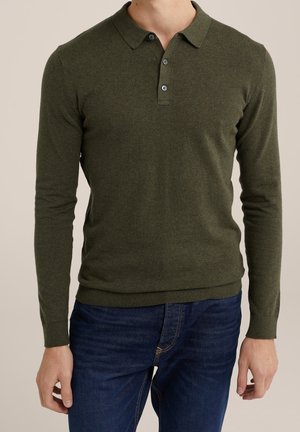 Mann trägt dunkelgrünen Polo-Pullover mit langen Ärmeln und geknöpftem Kragen sowie blaue Jeans vor neutralem Hintergrund.