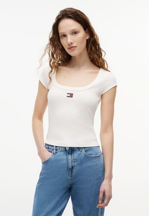 Νεαρή γυναίκα με σγουρά μαλλιά που φοράει λευκό εφαρμοστό τοπ με στρογγυλή λαιμόκοψη και μικρό λογότυπο, καθώς και ψηλόμεσα μπλε τζιν, με τα χέρια στις τσέπες.
