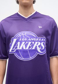 Άτομο που φοράει μοβ μπλούζα μπάσκετ των Los Angeles Lakers με λευκό λογότυπο και ύφασμα διχτυού σε απλό φόντο.