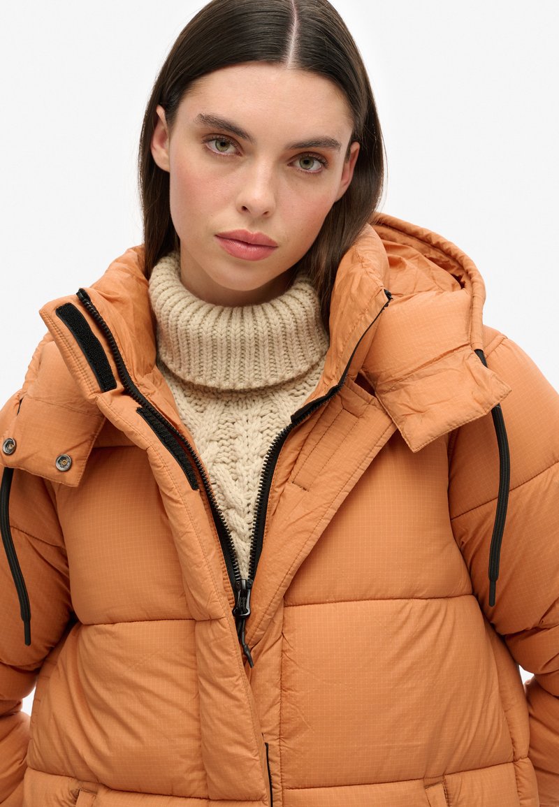 Gevoerde pufferjack in verbrand oranje met een ritsluiting, afneembare capuchon en zwarte accenten. Draag het over een chunky crèmekleurige trui.