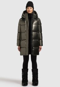 Padded coat v tmavozelenej, lesklej látke s kapucňou, dlhými rukávmi a rovnými líniami. Nosený cez čierny outfit a doplnený čiernymi topánkami.