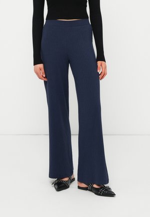 Femme portant un pantalon large côtelé bleu marine, un haut noir à manches longues et des ballerines slingback noires à bouts pointus avec des clous.
