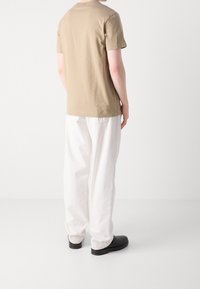 Beige bomulls-T-shirt med korta ärmar och rund halsringning, bärs tillsammans med vita, löst sittande byxor och svarta skor, sedd bakifrån.