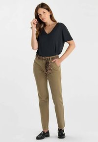 T-shirt noir à col en V associé à un pantalon kaki avec une ceinture nouée à imprimé léopard. Le mannequin pose avec une main sur la hanche, les chaussures sont noires.