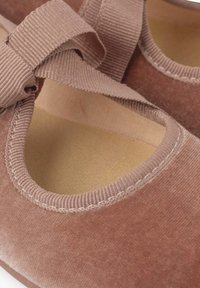 Scarpe in tessuto marrone con una consistenza morbida, caratterizzate da un'apertura arrotondata e una fascia a nastro abbinata sopra l'arco del piede. Solettini chiari visibili.