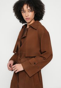 Barna szarvasbőr rövidített dzseki inggallérral és nagy első zsebekkel. Fekete gombokkal és sima textúrával rendelkezik.