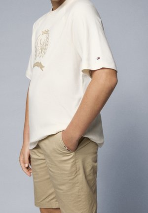 T-shirt beige à manches courtes avec un design de blason doré, associé à un short beige clair. Avec le logo Tommy Hilfiger sur la manche.
