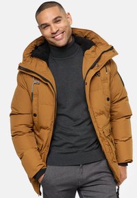 Brun puffervest med en hætte foret med pels, frontlommer og lynlås, båret over en mørk turtleneck. Med kontrasterende lynlåse.