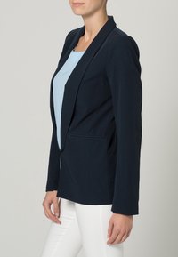 Marinblå blazer tillverkad av ett slätt tyg, med en skräddarsydd passform, slagkragar och två framfickor. Stylad med vita byxor.