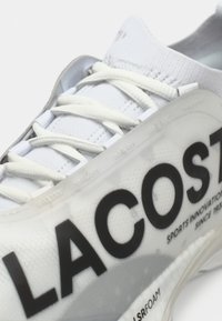Fehér Lacoste sportcipő, texturált hálós felsőrésszel. Jellemzője a feltűnő fekete logó és a könnyű talp sima elemekkel.