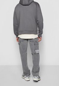 Felpa grigia con vestibilità rilassata, abbinata a pantaloni cargo grigi con una toppa logo bianca. L'outfit è completato da sneaker con texture.