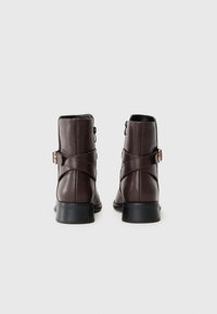 Botas de tobillo de cuero marrón con acabado mate, que cuentan con una correa ajustable y un tacón bajo en bloque. Cremallera en la parte trasera para un uso fácil.