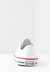 Białe trampki na płaskiej podeszwie, z niskim profilem, czerwono-niebieską paskowaną podeszwą oraz etykietą z logo na pięcie z napisem "Converse All Star."
