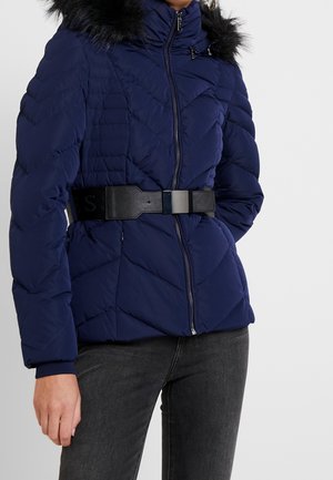 Modèle portant une doudoune matelassée bleu marine avec une capuche en fausse fourrure noire et une large ceinture, associée à un jean foncé.